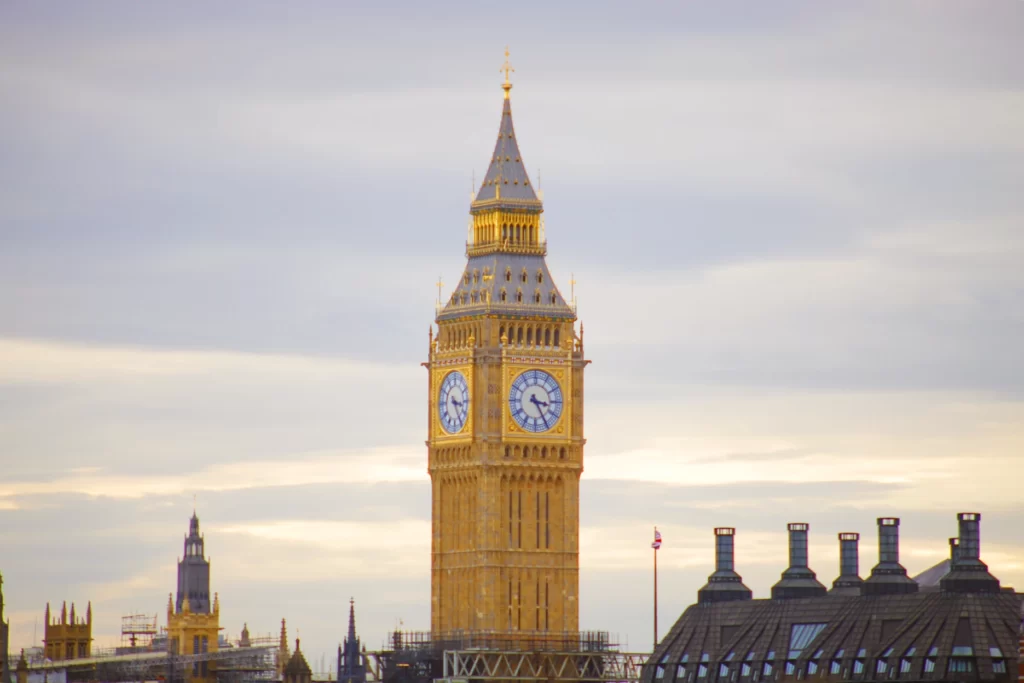 Елизаветинская башня или Биг-Бен в Лондоне. Вид на фоне крыш с шестью трубами и облачного неба. Часы показывают 15:25. Золотистая башня после ремонта сияет.
