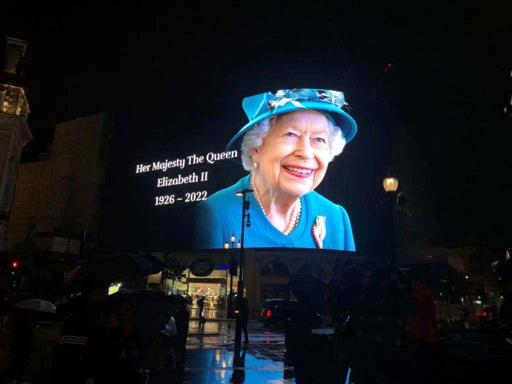 Бывшая Королева Великобритании, Елизавета Вторая на светодиодном экране на площади Пикадилли в Лондоне. Дождливая ночь. Все в черном цвете, так как Королева только что умерла и это в дань памяти. 