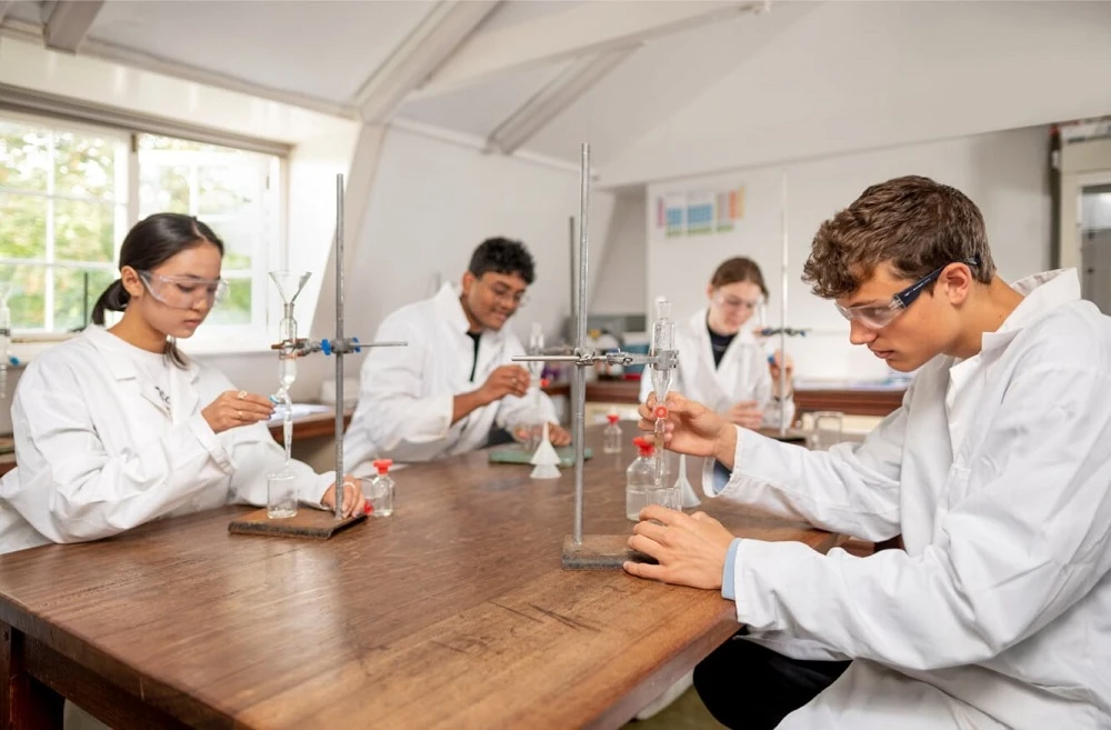 Школьники в английской школе, в белых халатах занимаются в лаборатории химией. Проводят эксперименты с реагентами.